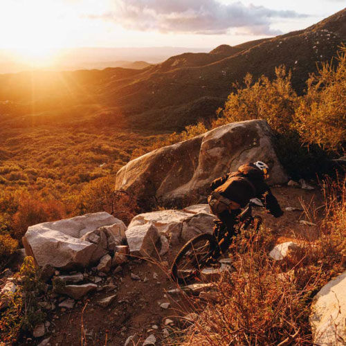 Rear view of a mountain biker riding through a rocky landscape with a sunset in the background.